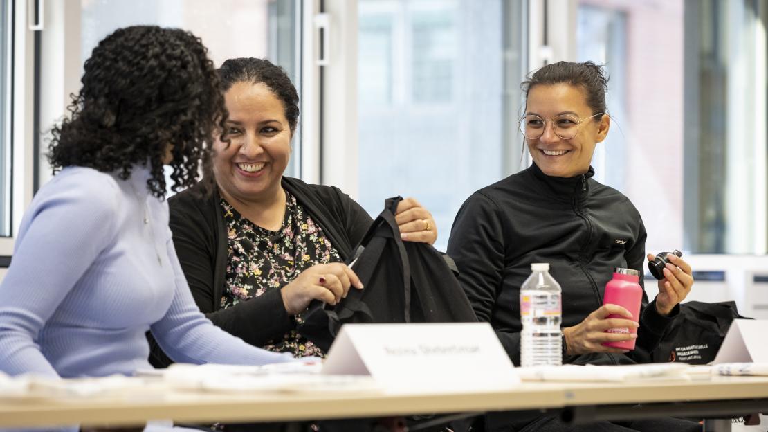 Drei Frauen lächelnd sitzend an Tischen in einem Seminarraum