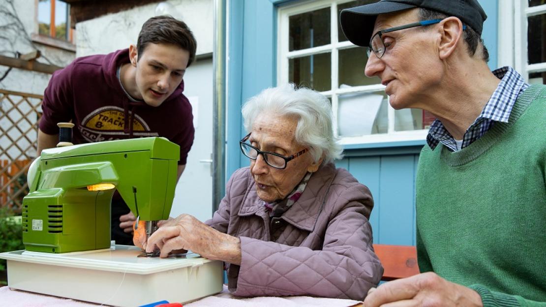 Eine ältere Dame arbeitet im Repair Café des Nachbarschaftszentrums Ginnheim an einer Nähmaschine, während zwei Männer ihr gespannt zusehen. 