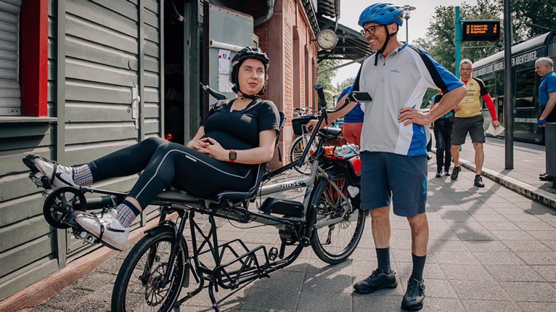 Eine sehbehinderte Frau sitzend auf einem Liegerad für zwei Personen unterhält sich mit einem Mann in Radlerkleidung, der anschließend mit ihr auf dem Rad fährt.