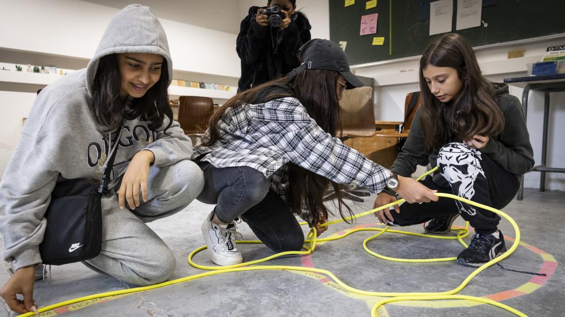 Mehrere Mädchen kniend auf dem Boden, die im Zuge eines Film-Workshops eine Art gelbes Kabel auf dem Boden auslegen.